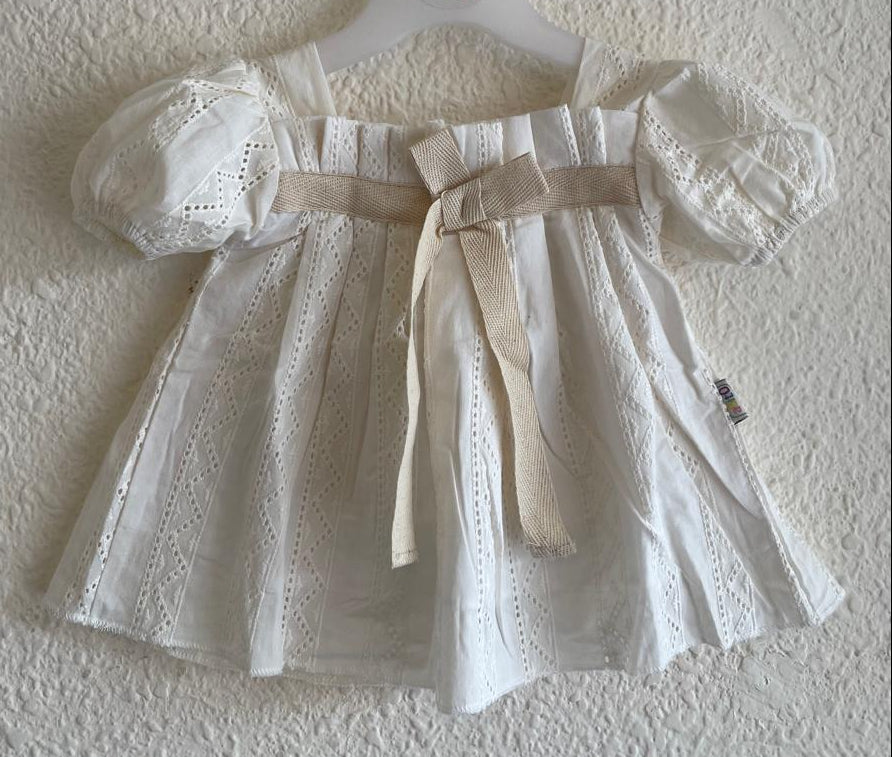 A white eyelet baby dress with puff sleeves and a beige ribbon bow at the waist, hanging on a white hanger against a textured white wall.