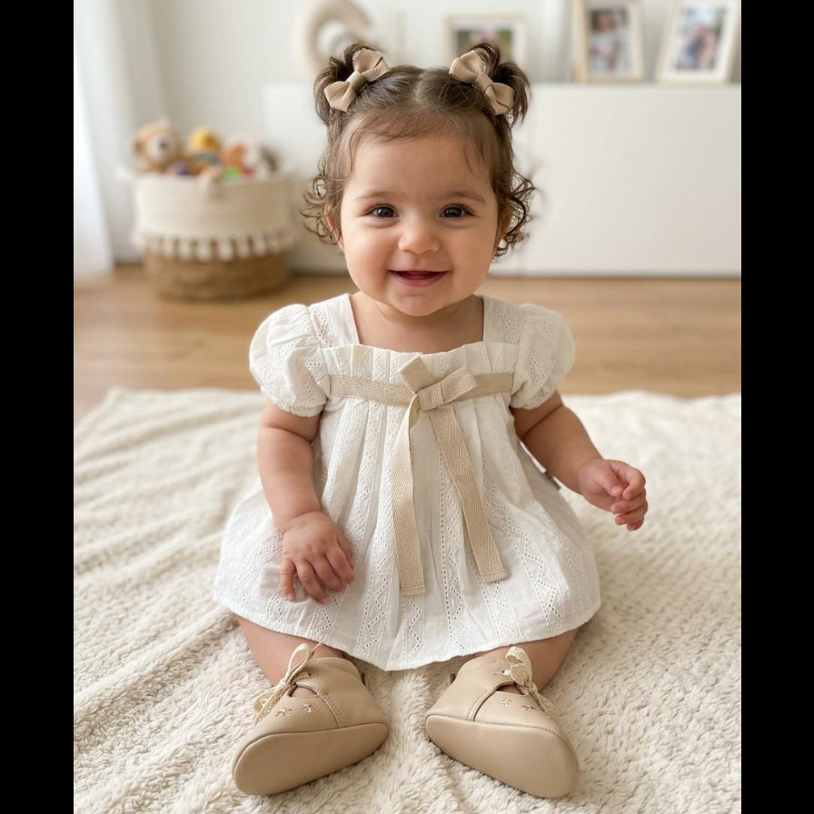 A smiling baby girl sitting on a cream textured rug, wearing a short-sleeve white eyelet dress with a beige ribbon bow at the waist and matching beige shoes. She has her hair in two buns with beige bows.