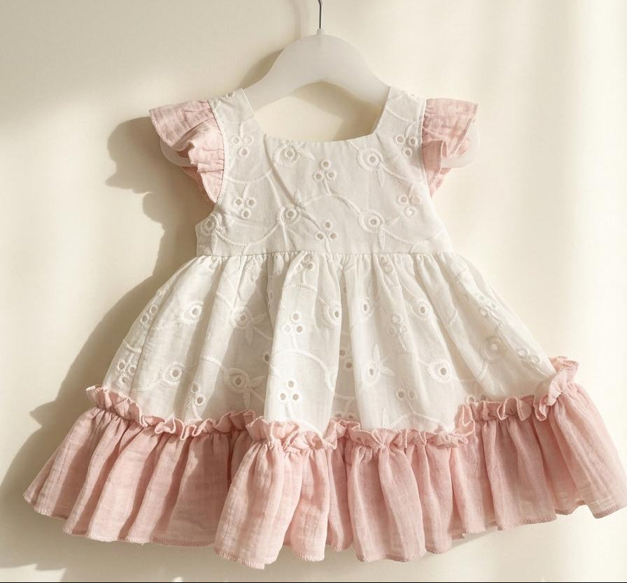 A baby girl's dress with a white eyelet bodice and a pink ruffled skirt, hanging on a white hanger against a warm beige wall.