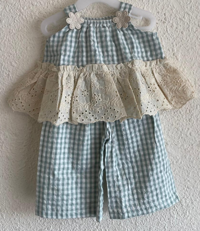 A baby girl's two-piece romper set hanging on a white plastic hanger against a textured white wall. The set features a light blue and white gingham check pattern on the top and shorts, with a beige eyelet lace ruffle layer across the chest.